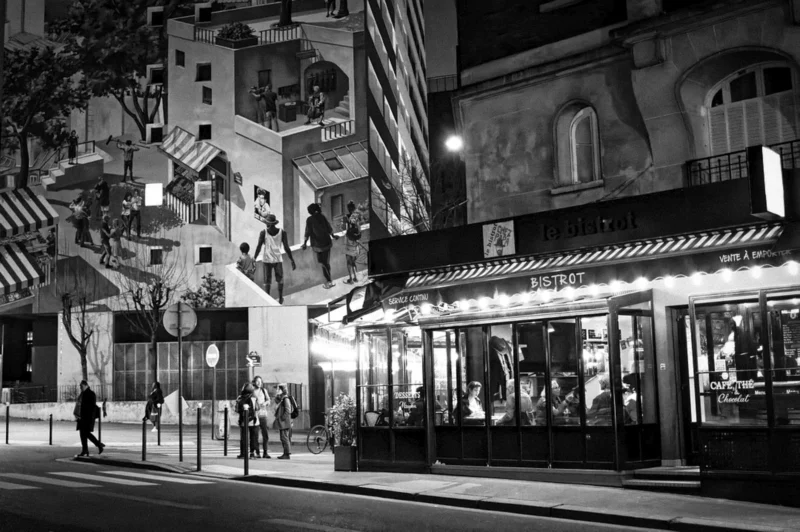 Thierry · 26/01/2024 · 3840 x 2553 Cette photographie en noir et blanc capture une rue nocturne animée. Le sujet principal semble être un bâtiment avec une façade ornée de fresques murales géométriques et des silhouettes humaines. La composition joue sur les contrastes entre les formes architecturales et les ombres. Le cadrage est serré sur le bâtiment, avec une perspective légèrement inclinée.La lumière, tamisée et artificielle, met en valeur les contours et crée une ambiance mystérieuse. Les couleurs dominantes, bien que monochromes, varient entre les tons clairs et sombres, renforçant le côté dramatique. Le style est urbain et artistique, évoquant une ambiance nocturne vivante et légèrement mélancolique.