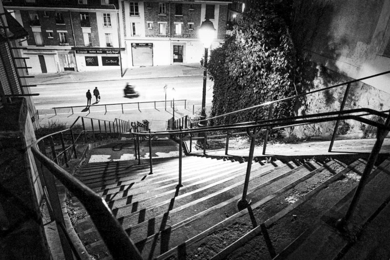 Thierry · 16/01/2024 · 3840 x 2560 Cette photographie en noir et blanc capture une scène urbaine nocturne. Le **sujet principal** est un escalier métallique en colimaçon descendant vers une rue déserte, éclairée par des réverbères.La **composition** est dynamique, avec une perspective descendante qui guide le regard. Le **cadrage** est serré, centré sur l’escalier et son environnement immédiat. La **lumière** est douce et contrastée, créant des ombres marquées.Les **couleurs dominantes** sont les nuances de gris, renforçant l’atmosphère nocturne. Le **style** est documentaire et esthétique, évoquant une ambiance **mélancolique et mystérieuse**, presque onirique.