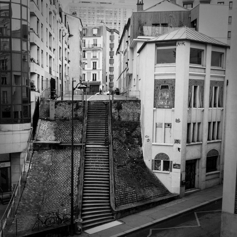 Thierry · 09/02/2024 · 3840 x 3840 Cette photographie en noir et blanc capture un escalier en pierre étroit encadré par des bâtiments urbains. Le sujet principal est cet escalier descendant entre des immeubles aux façades classiques. La composition est symétrique, avec des lignes directrices convergeant vers le bas de l’escalier.Le cadrage est serré, mettant en valeur les détails architecturaux. La lumière est naturelle, contrastée, créant des ombres marquées. Les couleurs dominantes sont les nuances de gris, renforçant l’aspect dramatique et historique. Le style est documentaire et urbain, évoquant une ambiance mélancolique et intemporelle.