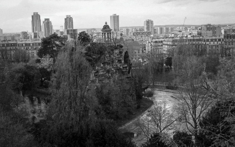 Thierry · 23/02/2024 · 3840 x 2400 Cette photographie en noir et blanc montre un parc urbain en hiver. Le sujet principal est un espace vert avec des arbres dénudés et des sentiers.La composition met en valeur un premier plan boisé et un arrière-plan de bâtiments, créant une profondeur. Le cadrage est large, englobant une vue panoramique.La lumière semble naturelle, mais diffuse, typique d’un jour couvert. Les couleurs dominantes sont les nuances de gris, accentuant les contrastes entre ombres et lumières.Le style est documentaire et sobre, évoquant une ambiance calme et mélancolique.