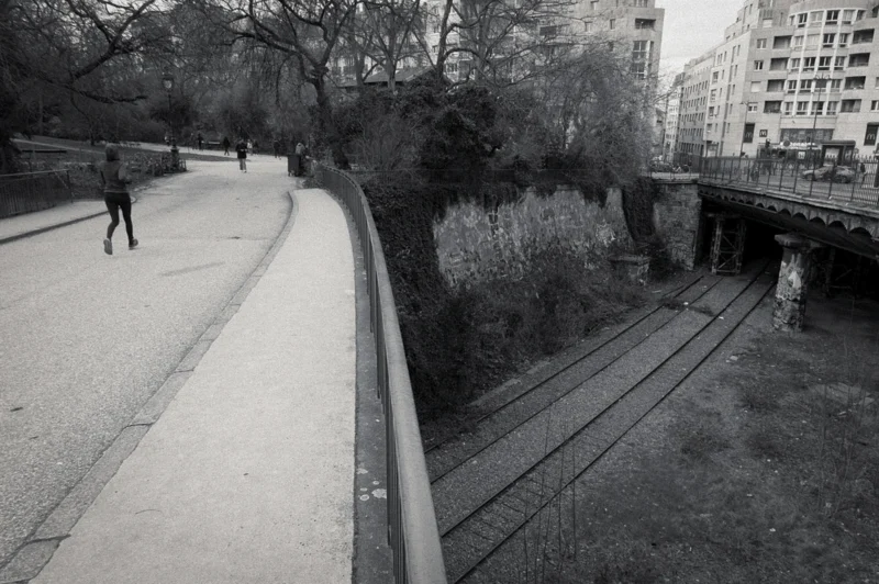 Thierry · 24/02/2024 · 3840 x 2553 Cette photographie en noir et blanc capture une scène urbaine solitaire. Le **sujet principal** est un **sentier piéton** bordant un **cours d’eau asséché**, avec une personne marchant à gauche.La **composition** est dynamique, avec des lignes directrices (le chemin et les rails) convergeant vers l’arrière-plan. Le **cadrage** est légèrement en contre-plongée, offrant une perspective immersive.La **lumière** est douce, sans contraste marqué, créant une ambiance **sombre et mélancolique**. Les **couleurs dominantes** sont les nuances de gris, renforçant l’aspect monochrome et intemporel.Le **style** est documentaire et minimaliste, évoquant une **ambiance calme et presque déserte**, typique des villes modernes.