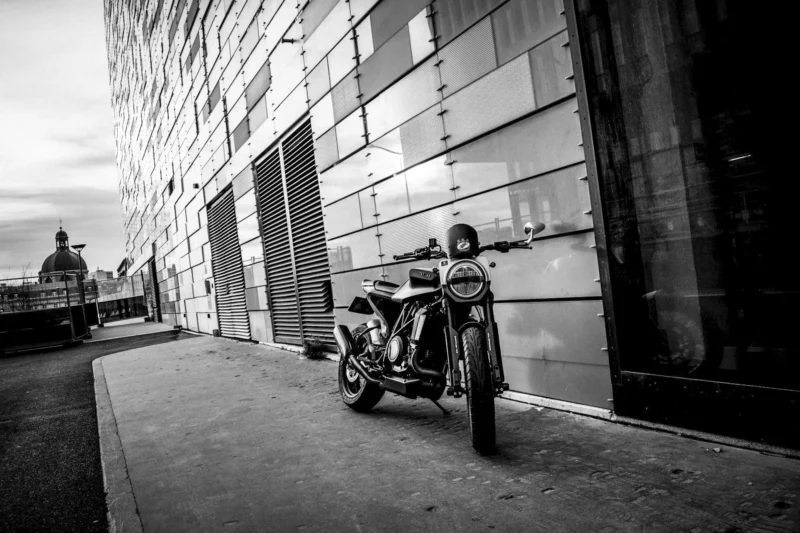 Thierry · 27/12/2023 · 3840 x 2559 Cette photographie en noir et blanc met en scène un **moto vintage** comme sujet principal, garé le long d’un trottoir urbain. La **composition** est simple et épurée, avec une perspective légèrement basse, accentuant la présence du véhicule.Le **cadrage** est serré, isolant la moto et les bâtiments modernes en arrière-plan. La **lumière**, douce et contrastée, crée des ombres marquées, renforçant l’ambiance dramatique. Les **couleurs dominantes** sont les nuances de gris, typiques du noir et blanc, avec des reflets plus clairs sur la moto.Le **style** est documentaire et esthétique, évoquant une **ambiance mélancolique et intemporelle**, mêlant modernité et nostalgie.