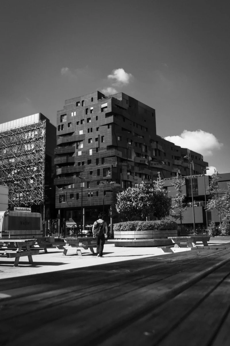 Thierry · 18/05/2024 · 2559 x 3840 Cette photographie en noir et blanc met en scène un environnement urbain. Le sujet principal est un immeuble moderne aux formes géométriques marquées, situé au centre. La composition joue sur les lignes architecturales et les perspectives, avec des bâtiments en arrière-plan et une rue en premier plan.Le cadrage inclut une portion de trottoir et des passants, ajoutant une touche de vie quotidienne. La lumière, naturelle et contrastée, accentue les ombres et les volumes. Les couleurs dominantes sont les nuances de gris, renforçant l’aspect dramatique et intemporel. Le style est documentaire et urbain, évoquant une ambiance à la fois dynamique et mélancolique.