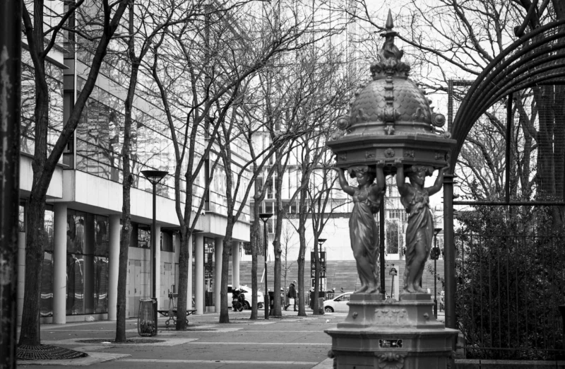 Thierry · 27/12/2023 · 3840 x 2507 Cette photographie en noir et blanc met en scène un **sujet principal** : une fontaine ornée, centrale et détaillée, entourée de barrières. La **composition** est symétrique, avec des arbres dénudés bordant une allée menant vers l’arrière-plan.Le **cadrage** est serré sur la fontaine, mais inclut aussi des bâtiments et des lampadaires. La **lumière**, douce et diffuse, crée des contrastes modérés, accentuant les textures. Les **couleurs dominantes** sont les gris et noirs, typiques du noir et blanc, avec des touches de blanc pour les éléments architecturaux.Le **style** est documentaire et classique, évoquant une **ambiance** calme et intemporelle, presque mélancolique.