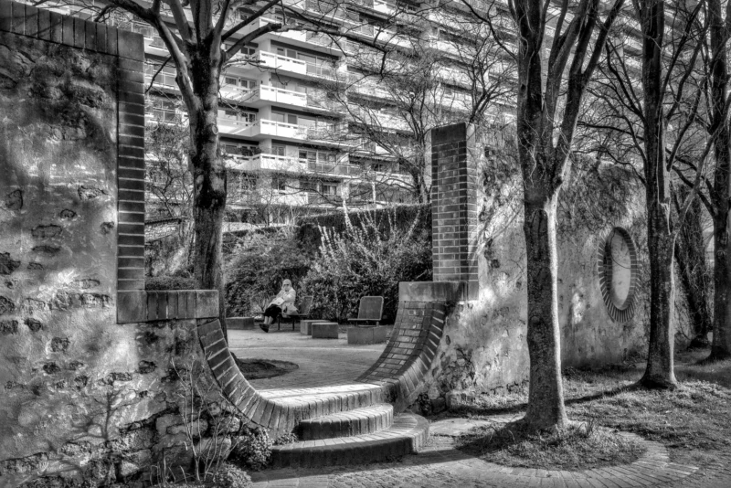 Thierry · 19/03/2024 · 3840 x 2561 Cette photographie en noir et blanc met en scène un **passage étroit entre deux bâtiments anciens**, encadré par des arbres dénudés. Le **sujet principal** est l’architecture, avec ses murs en briques et ses escaliers en pierre.La **composition** joue sur la perspective, créant un effet de profondeur et de mystère. Le **cadrage** serré isole le passage, renforçant l’intimité du lieu.La **lumière** douce et diffuse, probablement naturelle, accentue les textures et les ombres, ajoutant une atmosphère mélancolique. Les **tons de gris** dominent, soulignant les contrastes.Le style **sombre et contemplatif** évoque une ambiance **nostalgique et paisible**, presque onirique.
