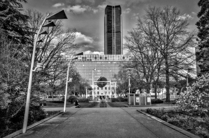 Thierry · 04/07/2025 · 3840 x 2528 Cette photographie en noir et blanc met en scène un grand bâtiment moderne, probablement un parlement ou une institution gouvernementale, avec une haute tour centrale. La composition est symétrique, encadrée par des arbres et des lampadaires de part et d’autre d’une allée menant vers l’édifice. Le cadrage est large, incluant un ciel partiellement nuageux.La lumière semble naturelle, avec des contrastes marqués entre ombres et clartés. Les couleurs dominantes, bien que monochromes, jouent sur les nuances de gris et de noir, renforçant l’ambiance solennelle et majestueuse du lieu. Le style est documentaire et classique, évoquant une atmosphère à la fois imposante et sereine.