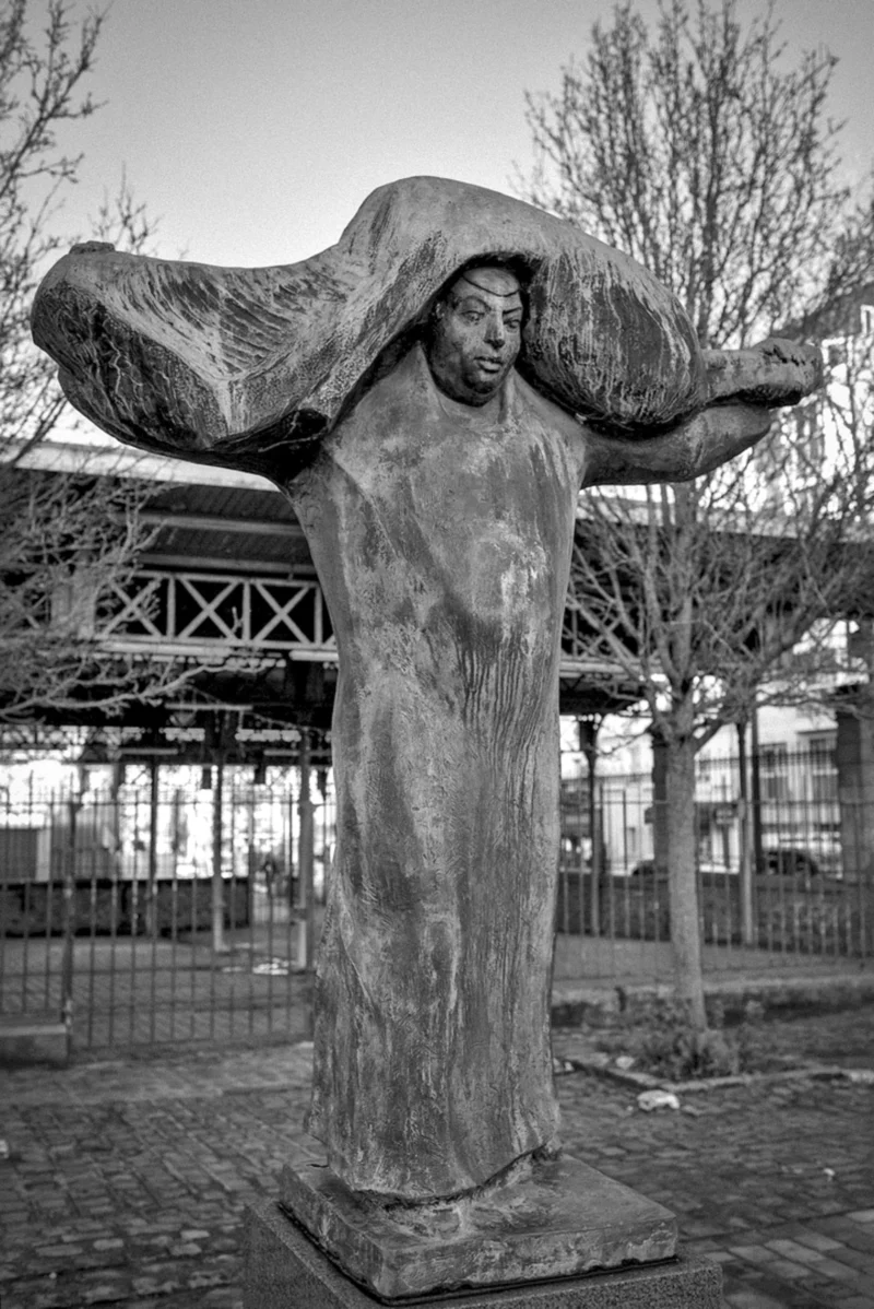 Thierry · 18/01/2024 · 2564 x 3840 Cette photographie montre une statue en bronze comme sujet principal, représentant une figure humaine aux bras levés, comme en mouvement. La composition est centrée sur la sculpture, entourée d’un environnement urbain avec des arbres et une structure métallique en arrière-plan.Le cadrage est serré, mettant en valeur les détails de la statue. La lumière, naturelle et douce, crée des contrastes d'ombres et de clartés. En noir et blanc, les tons varient du clair au foncé, accentuant les textures et les formes. L’ambiance est à la fois artistique et contemplative, évoquant une certaine mélancolie et puissance sculpturale.