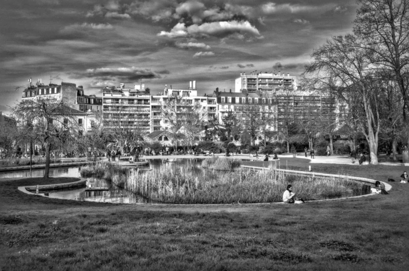 Thierry · 04/07/2025 · 3840 x 2542 Cette photographie en noir et blanc met en scène un parc urbain avec un étang central comme sujet principal. La composition est équilibrée, avec des bâtiments en arrière-plan et une végétation structurée autour de l’eau.Le cadrage est large, incluant un horizon dégagé. La lumière, douce et diffuse, crée des contrastes subtils. Les tons dominants varient du clair au foncé, renforçant la profondeur.Le style est documentaire et artistique, évoquant une ambiance calme et intemporelle, presque mélancolique, malgré l’absence de couleurs.
