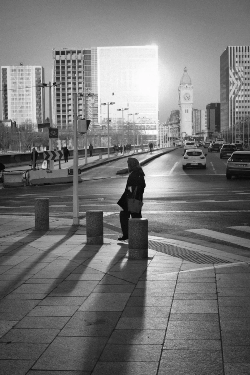 Thierry · 04/07/2025 · 2560 x 3840 Cette photographie en noir et blanc met en scène une femme comme sujet principal, debout près d’un poteau avec un sac, sur un trottoir urbain. La composition joue sur les lignes droites des pavés et des bâtiments, créant une perspective géométrique. Le cadrage inclut une avenue bordée de voitures et de gratte-ciels, avec une horloge emblématique en arrière-plan.La lumière est douce, avec des contrastes marqués entre ombres et clarté, accentuant l’ambiance urbaine. Les tons dominants sont les gris et noirs, renforçant une atmosphère moderne et légèrement mélancolique. Le style est épuré et documentaire, captant une scène de vie quotidienne.