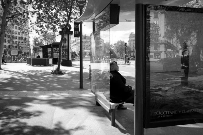 Thierry · 04/07/2025 · 3840 x 2560 Cette photographie en noir et blanc met en scène un homme assis seul sur un banc, sujet principal. La composition joue sur la symétrie avec des éléments urbains comme les affiches et les arbres. Le cadrage est serré sur le banc et l’homme, tout en intégrant l’environnement urbain en arrière-plan.La lumière est douce, créant des contrastes entre ombres et clartés. Les tons dominants sont les gris et noirs, renforçant une ambiance mélancolique et contemplative. Le style est documentaire et introspectif, évoquant une solitude urbaine.
