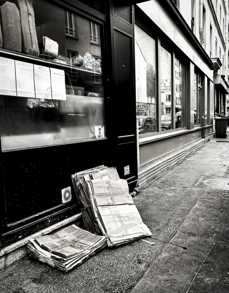 Thierry · 04/07/2025 · 3006 x 3840 Cette photographie en noir et blanc montre pour sujet principal un tas de journaux ou de papiers entassés contre un mur. La composition est simple et épurée, avec un cadrage serré sur les déchets et une partie de la vitrine d’un bâtiment en arrière-plan.La lumière est naturelle, probablement en journée, mais contrastée, créant des ombres marquées. Les couleurs dominantes sont les tons de gris, typiques du noir et blanc, renforçant une ambiance sobre et mélancolique. Le style est documentaire et réaliste, évoquant une atmosphère urbaine et négligée.