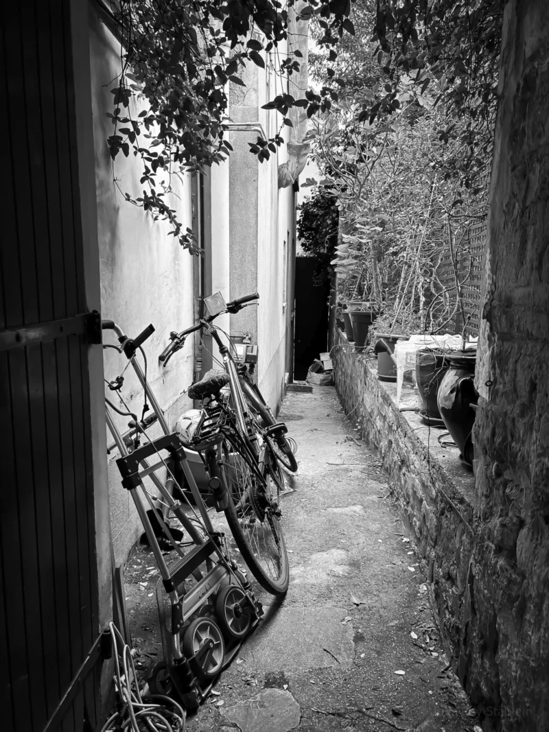 Thierry · 04/07/2025 · 2880 x 3840 Cette photographie en noir et blanc capture un **couloir étroit** bordé de murs et de végétation. Le **sujet principal** est un **vélo appuyé contre un mur**, partiellement caché par une porte entrouverte. La **composition** est simple et asymétrique, avec une perspective plongeante.Le **cadrage** est serré, guidant le regard vers le fond de l’allée. La **lumière** est naturelle, tamisée, créant des contrastes doux. Les **couleurs dominantes** sont les gris et noirs, accentuant l’atmosphère mélancolique et intimiste. Le **style** est documentaire et sobre, évoquant une **ambiance calme et légèrement nostalgique**.