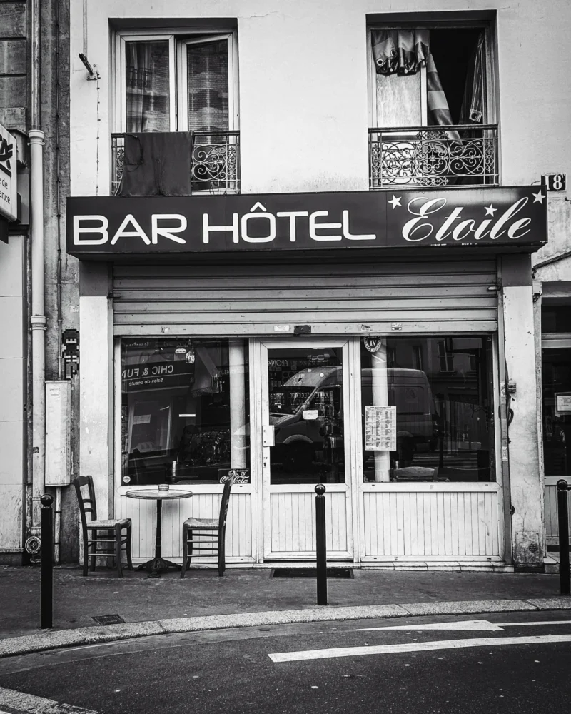 Thierry · 04/07/2025 · 3072 x 3840 Cette photographie en noir et blanc met en scène un **bar-hôtel nommé "Étoile"**. Le sujet principal est la façade du bâtiment, avec son enseigne lumineuse et ses fenêtres.La **composition** est symétrique, centrée sur l’entrée. Le **cadrage** est serré, capturant une partie de la rue et un trottoir. La **lumière**, contrastée, accentue les ombres et les détails architecturaux.Les **couleurs dominantes** sont les nuances de gris, renforçant l’ambiance rétro. Le style est **documentaire et vintage**, évoquant une atmosphère **nostalgique et urbaine**, typique des quartiers historiques.