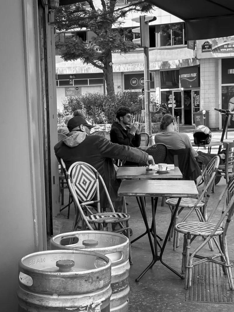 Thierry · 21/05/2024 · 2880 x 3840 Cette photographie en noir et blanc capture une scène intimiste dans un café extérieur. Le sujet principal est un groupe de personnes assises autour de tables, engagées dans une conversation. La composition met en avant les tables et chaises en métal, ainsi que des kegs de bière au premier plan.Le cadrage est serré, centré sur les convives et l’espace immédiat du café. La lumière est douce, probablement naturelle, créant des contrastes modérés. L’ambiance est calme et conviviale, évoquant une pause détente entre amis ou collègues. Le style est documentaire, réaliste, renforcé par le traitement monochrome.