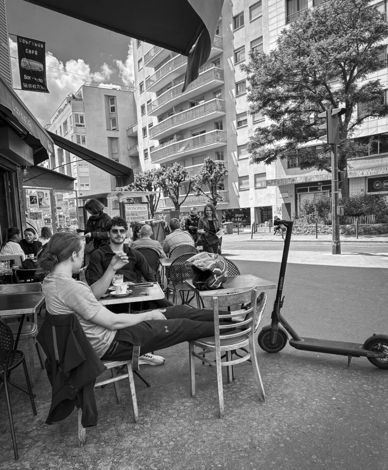 Thierry · 21/05/2024 · 3173 x 3840 Cette photographie en noir et blanc capture un groupe de personnes attablées dans un café en extérieur. Le sujet principal est une scène conviviale de sociabilité urbaine.La composition montre des tables disposées sur une terrasse, avec des bâtiments et des arbres en arrière-plan. Le cadrage inclut une partie de la rue et un trottinette électrique à droite. La lumière semble naturelle, probablement en journée, créant des contrastes doux.Les tons dominants sont les gris et noirs, typiques du noir et blanc, renforçant une ambiance à la fois moderne et intemporelle. Le style est documentaire et réaliste, évoquant une ambiance détendue et quotidienne.