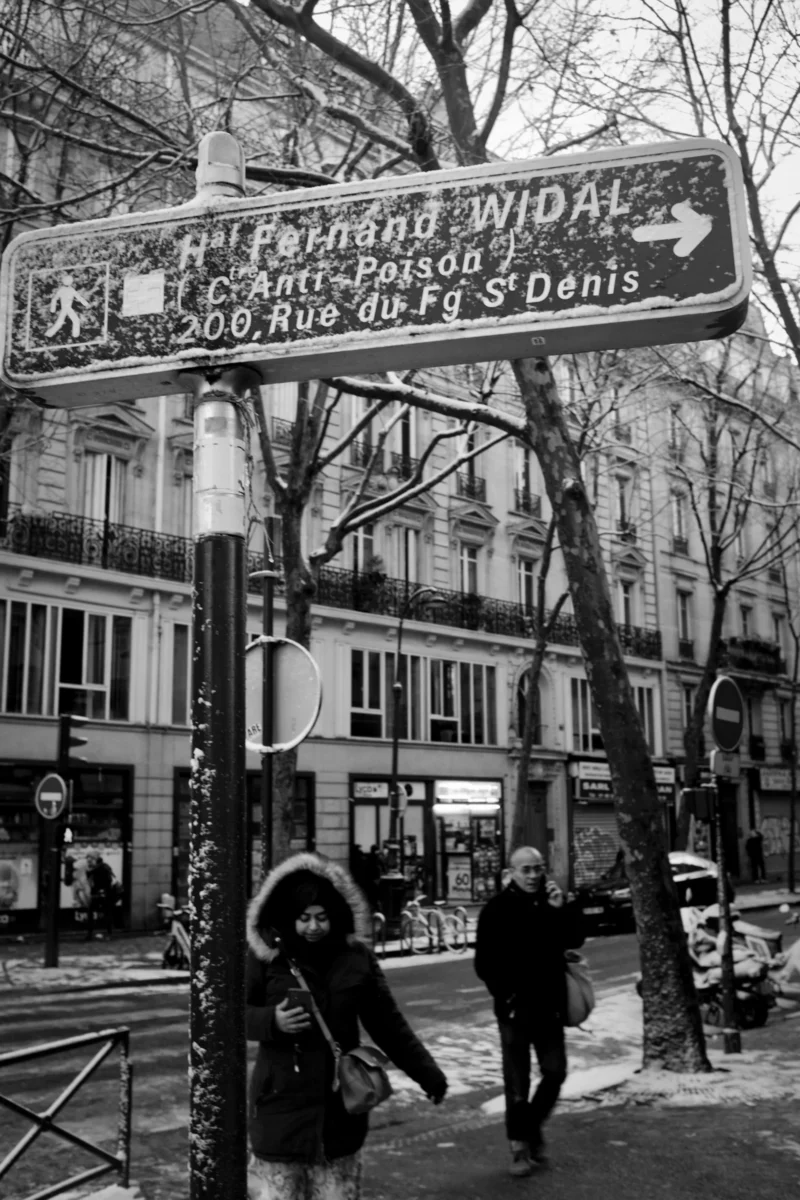 Thierry · 22/01/2014 · 2560 x 3840 Cette photographie en noir et blanc montre une rue parisienne avec une enseigne indiquant "Rue Pasteur" et "Rue Widal". Le sujet principal est un panneau de rue, avec deux personnes en mouvement en premier plan.La composition est équilibrée, avec le panneau central et les bâtiments en arrière-plan. Le cadrage est serré sur les détails urbains. La lumière est naturelle, probablement en journée, créant des contrastes marqués. Les couleurs dominantes, bien que monochromes, suggèrent des tons gris et noirs.Le style est documentaire, capturant une scène de vie quotidienne. L’ambiance est urbaine et authentique, évoquant le charme des rues parisiennes.