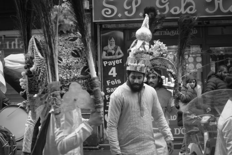 Thierry · 21/01/2024 · 3840 x 2560 Cette photographie en noir et blanc met en scène un homme comme sujet principal, vêtu d’un costume traditionnel et d’un chapeau orné. Il se tient devant une boutique aux enseignes colorées, partiellement visibles.La composition montre un cadrage serré, centré sur l’homme, avec des éléments décoratifs et des objets artisanaux autour de lui. La lumière semble naturelle, avec des contrastes marqués créant des ombres nettes. L’ambiance est à la fois traditionnelle et vivante, évoquant une scène culturelle ou festive dans un marché ou une rue animée. Le style est documentaire et artistique, captant un moment authentique.