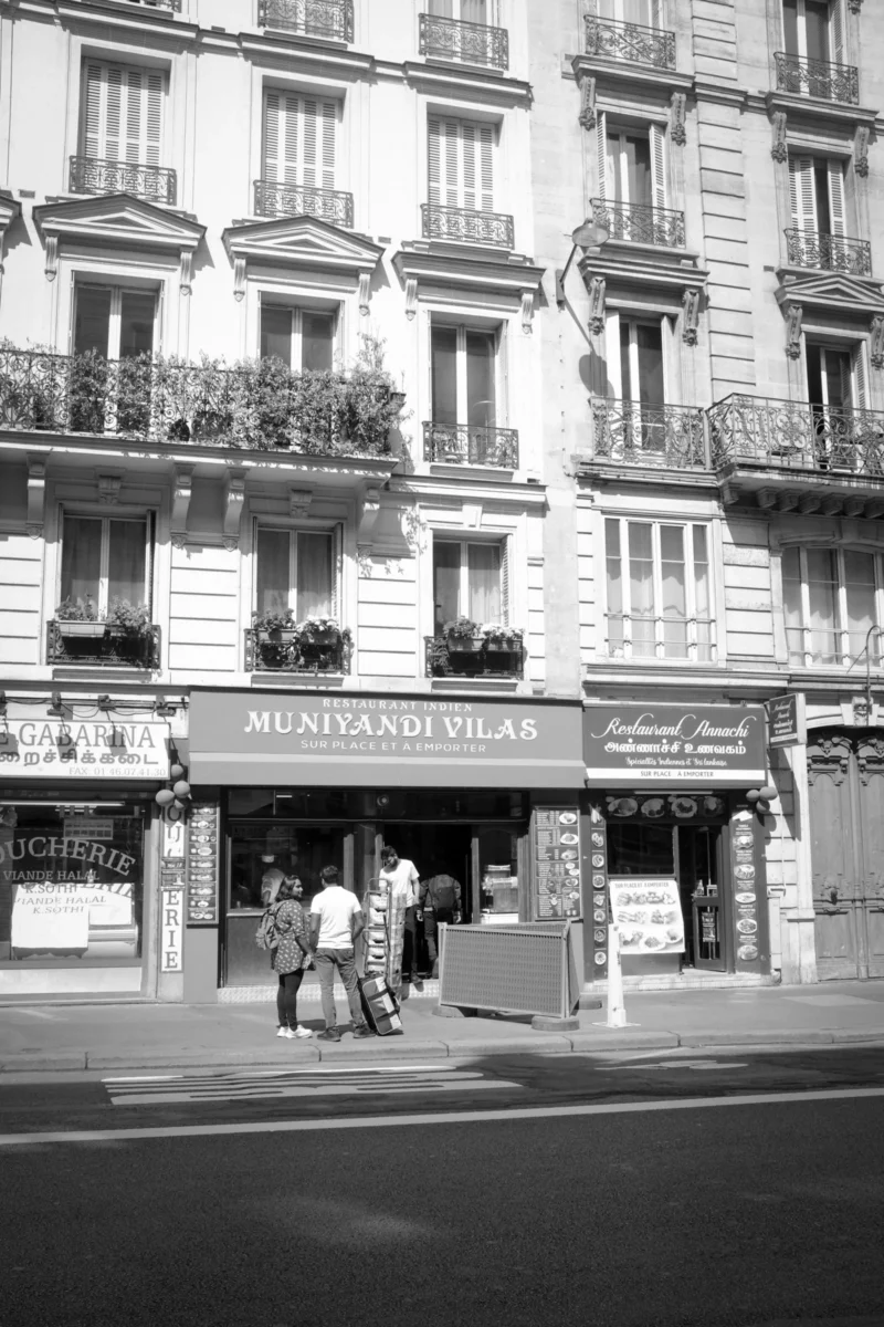 Thierry · 25/05/2014 · 2560 x 3840 Cette photographie en noir et blanc capture une façade parisienne typique. Le sujet principal est une boutique nommée "Montand & Pilar", entourée de balcons fleuris. La composition montre une rue animée avec des passants et des bâtiments classiques.Le cadrage est serré sur la boutique et les immeubles environnants. La lumière est naturelle, avec des contrastes marqués entre ombres et clartés. Les couleurs dominantes, bien que monochromes, évoquent des tons gris et blancs.Le style est documentaire, rappelant les clichés des rues parisiennes des années 1950-1970. L’ambiance est calme et rétro, empreinte d’un charme intemporel.