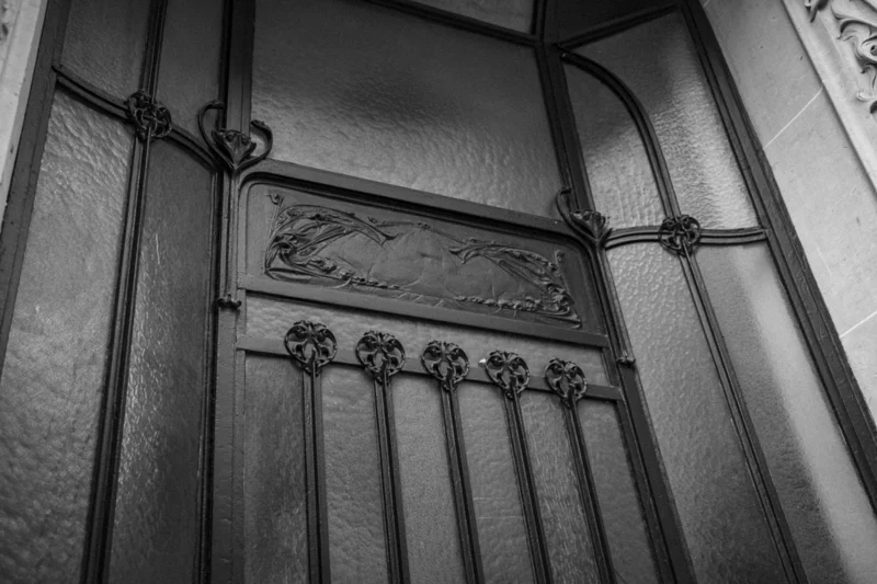 Thierry · 04/07/2025 · 3840 x 2560 Cette photographie met en scène une porte ancienne en fer forgé. Le sujet principal est le panneau central orné d’un motif décoratif stylisé, probablement une lettre ou un emblème.La composition est symétrique, avec des barres verticales encadrant le panneau. Le cadrage est serré, mettant l’accent sur les détails.La lumière, douce et tamisée, crée des contrastes nets entre les zones éclairées et ombragées. Les couleurs dominantes sont les tons de gris et noir, typiques d’une photo en noir et blanc.Le style est classique et élégant, évoquant une ambiance historique et mystérieuse.