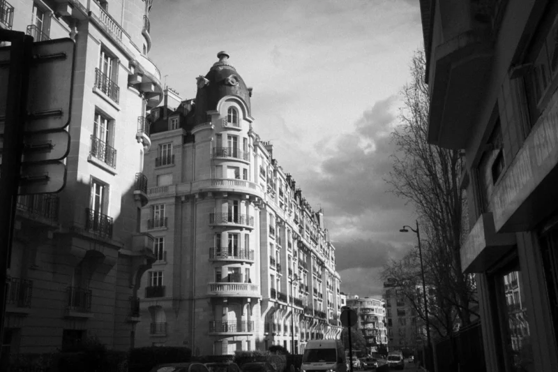 Thierry · 04/07/2025 · 3840 x 2560 Cette photographie en noir et blanc capture une rue urbaine bordée d'immeubles anciens. Le sujet principal est l’architecture des bâtiments, avec leurs façades ornées et leurs balcons en fer forgé. La composition est symétrique, avec une perspective convergente vers le lointain.Le cadrage est serré, mettant en valeur les détails architecturaux. La lumière est tamisée, créant des contrastes entre ombres et clartés, renforçant l'atmosphère mélancolique. Les tons dominants sont les gris et noirs, typiques du monochrome.Le style évoque un réalisme documentaire, tandis que l’ambiance, à la fois calme et nostalgique, rappelle les quartiers historiques des grandes villes européennes.
