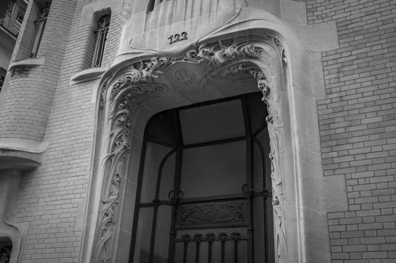 Thierry · 04/07/2025 · 3840 x 2560 Cette photographie met en scène une entrée d’immeuble ornée d’un style architectural Art nouveau. Le sujet principal est l’arche décorative au-dessus de la porte, richement sculptée de motifs végétaux. La composition est symétrique, centrée sur l’entrée numérotée 322.Le cadrage est serré, mettant en valeur les détails de la porte en fer forgé et de l’archivolte. La lumière, douce et naturelle, crée des contrastes nets entre ombres et lumières, accentuant les textures. En noir et blanc, les tons dominants varient du blanc au gris foncé.L’ambiance est à la fois élégante et mystérieuse, évoquant un charme ancien et intemporel.
