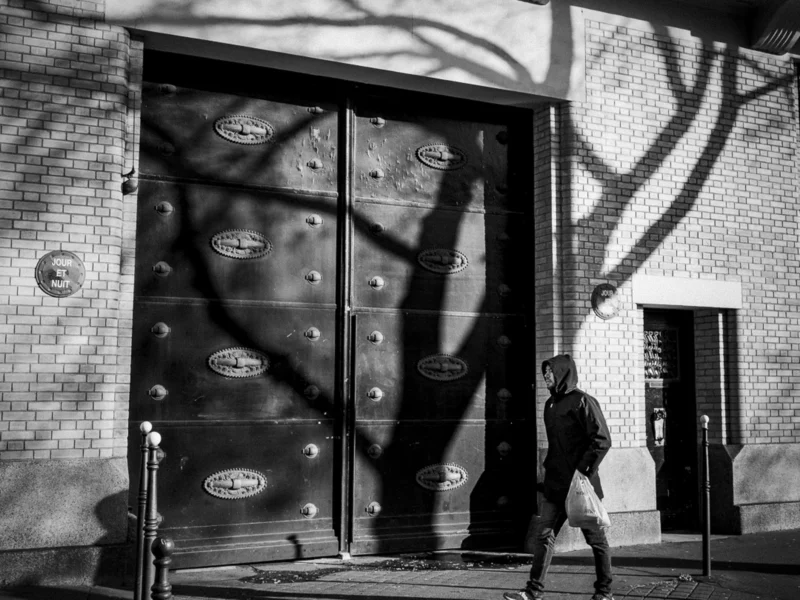 Thierry · 04/07/2025 · 3840 x 2880 Cette photographie en noir et blanc met en scène un homme marchant devant une porte métallique ornée de motifs en relief évoquant des muscles. Le sujet principal est cet homme, dont la silhouette est partiellement éclairée.La composition joue sur les contrastes entre les ombres et la lumière, créant une ambiance mystérieuse. Le cadrage serré et l'angle légèrement bas accentuent la profondeur et le dynamisme. Les jeux d'ombres et de lumières, ainsi que les textures des murs et de la porte, renforcent le style urbain et industriel. L'ambiance est à la fois sombre et intrigante, avec une touche de mélancolie.