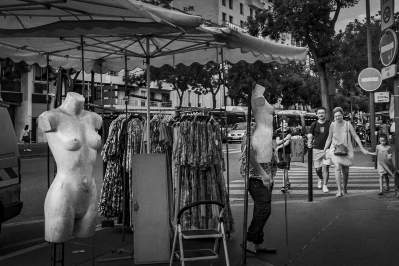 Thierry · 04/07/2025 · 3840 x 2560 Cette photographie en noir et blanc capture un marché en plein air. Le sujet principal est un mannequin vêtu de robes exposées sur des portants, entourés de vêtements suspendus. La composition montre des étals de vêtements et des passants en arrière-plan, créant une ambiance animée.Le cadrage est serré sur les vêtements et le mannequin, tout en incluant des éléments périphériques. La lumière est naturelle, douce, avec des contrastes modérés. Les couleurs, bien que monochromes, suggèrent des tons clairs et sombres typiques d’un marché.Le style est documentaire, réaliste, évoquant une scène de vie quotidienne. L’ambiance est vivante et populaire.