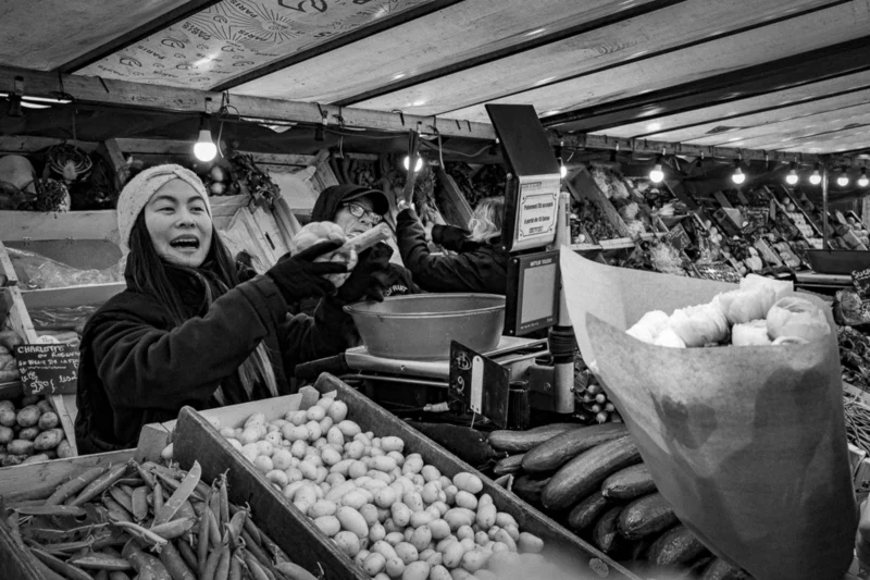 Thierry · 04/07/2025 · 3840 x 2560 Cette photographie en noir et blanc capture une scène de marché nocturne. Le sujet principal est une vendeuse souriante, vêtue d’un bonnet, en train de servir des clients. La composition montre des étals remplis de produits, notamment des pommes de terre.Le cadrage est serré sur les personnes et les produits, créant une ambiance intime. La lumière, tamisée et artificielle, accentue les contrastes et donne une touche dramatique. Les couleurs dominantes, bien que monochromes, évoquent des tons de gris et noirs profonds.Le style est documentaire et réaliste, reflétant une ambiance chaleureuse et authentique.