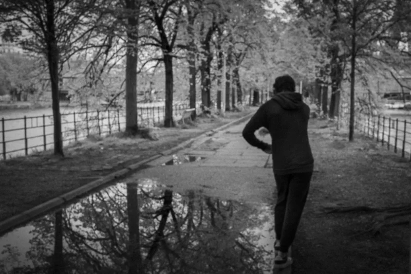 Thierry · 04/07/2025 · 3840 x 2560 Cette photographie en noir et blanc met en scène une personne marchant dos à la caméra le long d'un chemin bordé d'arbres. Le sujet principal est cette silhouette solitaire, renforcée par le cadrage serré et la perspective.La composition utilise une ligne centrale menant vers l'horizon, avec une flaque d'eau reflétant partiellement l'arbre, ajoutant une symétrie subtile. La lumière douce et diffuse crée une ambiance contemplative et mélancolique. Les tons dominants, variant du clair au foncé, accentuent la profondeur et le mystère. Le style sobre et épuré confère une touche intemporelle et poétique à l'image.
