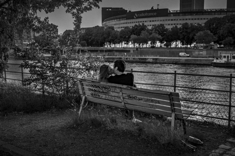 Thierry · 04/07/2025 · 3840 x 2560 Cette photographie en noir et blanc met en scène un couple assis sur un banc, dos à nous, observant un canal. Le sujet principal est leur posture intimiste et contemplative. La composition est équilibrée, avec le banc en premier plan et les bâtiments en arrière-plan, encadrés par des arbres.Le cadrage serré isole le couple tout en intégrant l’environnement urbain. La lumière douce, probablement crépusculaire, adoucit les contours et crée une ambiance mélancolique. Les tons dominants sont les gris et noirs, renforçant une atmosphère nostalgique et introspective. Le style est documentaire et poétique.