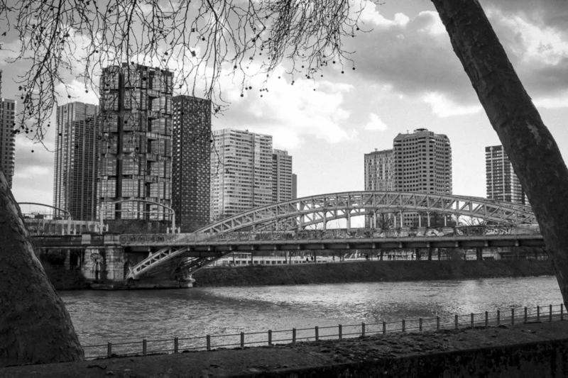 Thierry · 04/07/2025 · 3840 x 2560 Cette photographie en noir et blanc met en scène un **pont métallique** comme sujet principal, traversant un cours d’eau. La **composition** joue sur les lignes architecturales des bâtiments modernes en arrière-plan et du pont, créant une perspective dynamique. Le **cadrage** inclut une branche d’arbre en premier plan, ajoutant une touche naturelle.La **lumière**, douce et diffuse, accentue les contrastes sans être trop forte. Les **couleurs dominantes** sont les nuances de gris, renforçant l’aspect urbain et moderne. Le **style** est documentaire et esthétique, évoquant une **ambiance** à la fois calme et industrielle, mêlant nature et urbanisation.