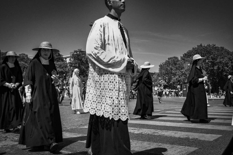 Thierry · 04/07/2025 · 3840 x 2560 Cette photographie en noir et blanc met en scène un homme vêtu d’une robe blanche et d’un chapeau large, semblant être le sujet principal. Il est entouré de plusieurs femmes en habits sombres, probablement des nonnes, avec des voiles et des coiffes traditionnelles.La composition est symétrique et centrée sur l’homme, créant un contraste visuel fort. Le cadrage est serré, mettant l’accent sur les personnages et leur interaction. La lumière est douce, avec des ombres marquées, renforçant l’atmosphère contemplative et solennelle. Le style est documentaire et intemporel, évoquant une scène religieuse ou culturelle. L’ambiance est calme et mystérieuse.