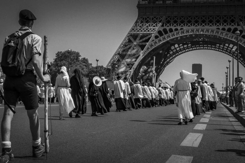 Thierry · 04/07/2025 · 3840 x 2560 Cette photographie met en scène une procession militaire sous l’arc de triomphe de l’Eiffel, à Paris. Le sujet principal est le défilé des soldats en uniformes traditionnels, encadrés par des spectateurs.La composition joue sur la perspective avec un cadrage serré sur les participants en premier plan et l’Eiffel en arrière-plan. La lumière est naturelle, douce, créant des contrastes nets en noir et blanc. Les couleurs dominantes, bien que monochromes, soulignent les ombres et les formes.Le style est documentaire et sobre, renforçant une ambiance solennelle et historique.
