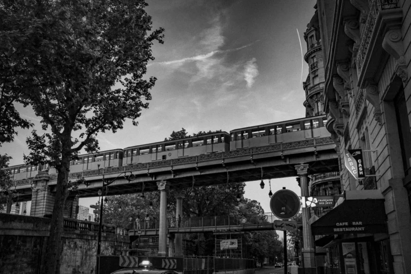 Thierry · 04/07/2025 · 3840 x 2560 Cette photographie en noir et blanc met en scène un **viaduc urbain** avec une structure métallique supportant une voie ferrée ou un tramway. Le **sujet principal** est ce pont surplombant une rue animée.La **composition** joue sur les lignes horizontales et verticales, créant une perspective dynamique. Le **cadrage** inclut des éléments urbains comme des arbres, des bâtiments et des panneaux routiers.La **lumière** est douce, avec des contrastes marqués, accentuant les ombres et les textures. Les **couleurs dominantes** sont les nuances de gris, renforçant l’aspect dramatique et intemporel.Le **style** est documentaire et esthétique, évoquant une **ambiance** à la fois moderne et nostalgique, typique des villes européennes.