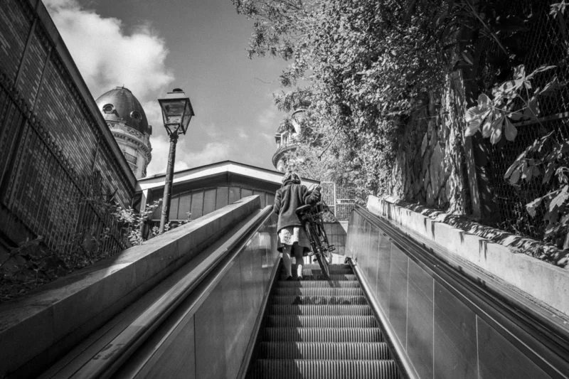 Thierry · 04/07/2025 · 3840 x 2560 Cette photographie en noir et blanc met en scène un escalier extérieur comme sujet principal. La composition est dynamique, avec des lignes convergentes menant vers le haut de l'escalier. Le cadrage est serré, capturant une personne en mouvement et une autre assise.La lumière, naturelle et contrastée, accentue les ombres et les textures. Les couleurs dominantes, bien que monochromes, varient du blanc au noir en passant par des gris.Le style est documentaire et artistique, évoquant une ambiance mélancolique et contemplative. Les jeux d'ombres et de lumière renforcent une atmosphère à la fois urbaine et intemporelle.
