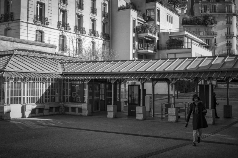 Thierry · 04/07/2025 · 3840 x 2560 Cette photographie en noir et blanc capture une scène urbaine calme. Le sujet principal est un marché couvert avec des étals vides sous une structure métallique. La composition met en valeur les bâtiments anciens en arrière-plan, avec des balcons et des fenêtres typiques. Le cadrage inclut une personne marchant à droite, ajoutant une touche de vie.La lumière est douce, probablement naturelle et tamisée, créant des contrastes subtils. Les couleurs dominantes, bien que monochromes, jouent sur les nuances de gris et de noir. Le style est documentaire et intemporel, évoquant une ambiance mélancolique et paisible, typique d'un quartier historique.