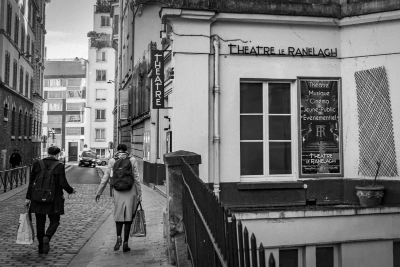 Thierry · 04/07/2025 · 3840 x 2560 Cette photographie en noir et blanc capture une scène urbaine. Le sujet principal est un couple marchant sur un trottoir en pente vers un théâtre nommé "Théâtre de l'Eldorado" (ici "Ranelagh" semble être une erreur). La composition est dynamique, avec une perspective légèrement inclinée et un cadrage serré sur les bâtiments et les passants.La lumière est douce, créant des contrastes subtils. Les couleurs dominantes, bien que monochromes, varient du noir au gris clair. Le style est documentaire et intemporel, évoquant une ambiance mélancolique et calme, typique des rues européennes.