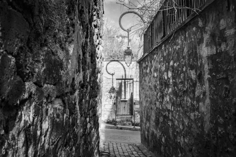 Thierry · 04/07/2025 · 3840 x 2560 Cette photographie en noir et blanc capture une ruelle étroite et ancienne. Le sujet principal est une porte en bois avec un heurtoir en forme d’anneau, encadrée par des murs de pierre usés.La composition joue sur la perspective profonde, avec un cadrage serré qui guide le regard vers l’arrière-plan. La lumière, tamisée et douce, accentue les textures et crée des contrastes entre ombres et clartés. Les couleurs dominantes, bien que monochromes, varient du gris clair au noir profond.Le style est documentaire et artistique, évoquant une ambiance mystérieuse et intemporelle, presque mélancolique.