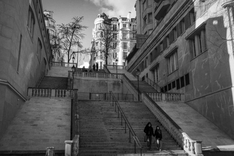Thierry · 04/07/2025 · 3840 x 2560 Cette photographie en noir et blanc capture un escalier urbain étroit encadré par des bâtiments. Le sujet principal est l’escalier, avec quelques personnes en mouvement, créant une impression de profondeur.La composition repose sur des lignes convergentes menant vers le haut, renforçant le dynamisme. Le cadrage est serré, mettant en valeur les détails architecturaux et les jeux d’ombres. La lumière, douce et contrastée, accentue les textures et les volumes.Les tons dominants varient du noir au gris clair, ajoutant une ambiance mélancolique et intemporelle. Le style est documentaire et réaliste, évoquant une atmosphère à la fois quotidienne et poétique.