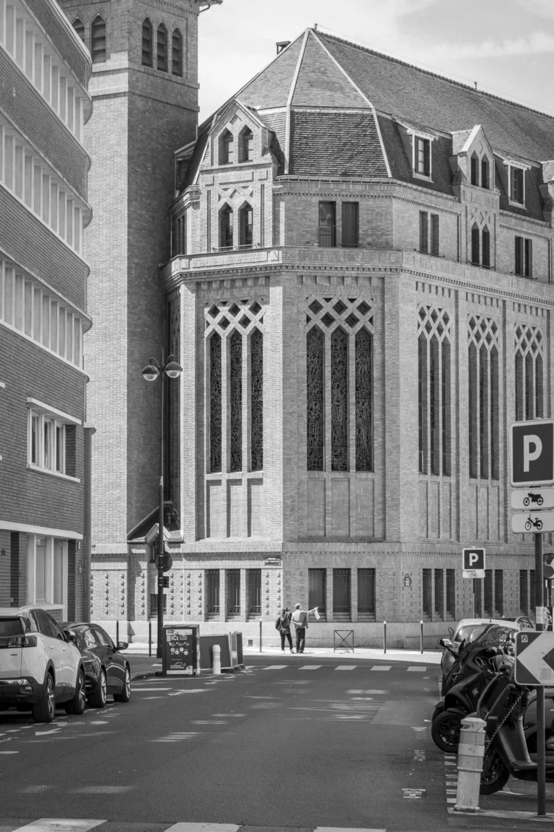 Thierry · 10/05/2024 · 2560 x 3840 Cette photographie en noir et blanc met en scène un bâtiment religieux de style gothique, avec des arcs brisés et des vitraux. Le sujet principal est cette église, dont la façade imposante domine l’image.La composition montre une rue urbaine avec des voitures et des piétons, encadrée par des immeubles modernes. Le cadrage inclut une perspective légèrement basse, accentuant la hauteur de l’édifice. La lumière semble naturelle, avec des contrastes marqués.L’ambiance est à la fois historique et contemporaine, mêlant tradition architecturale et vie urbaine moderne. Le monochrome renforce le côté intemporel de la scène.