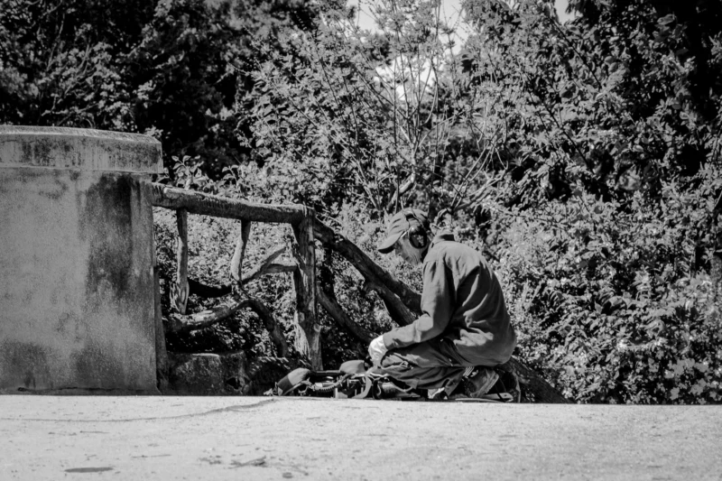 Thierry · 10/05/2024 · 3840 x 2560 Cette photographie en noir et blanc met en scène un homme assis seul sur un banc, le sujet principal. La composition repose sur une mise en valeur de sa solitude, entourée de végétation dense.Le cadrage est serré sur lui, avec un arrière-plan légèrement flou. La lumière, douce et naturelle, semble provenir de la gauche, créant des ombres légères. Les tons dominants sont les gris et noirs, renforçant une ambiance mélancolique et contemplative. Le style est documentaire et introspectif, évoquant une atmosphère calme et presque nostalgique.