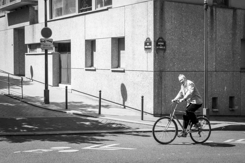 Thierry · 10/05/2024 · 3840 x 2560 Cette photographie en noir et blanc met en scène un cycliste comme sujet principal, vêtu d’un costume et d’un chapeau, en mouvement sur une route urbaine.La composition est dynamique, avec un cadrage serré sur le cycliste, centré dans l’image. L’arrière-plan montre un bâtiment et des éléments urbains comme des panneaux et des barrières.La lumière est naturelle, contrastée, accentuant les ombres et les formes. Les tons dominants sont les gris et noirs, typiques du monochrome, renforçant une ambiance rétro et intemporelle. Le style évoque un documentaire classique, évoquant une scène de vie quotidienne dans une ville.