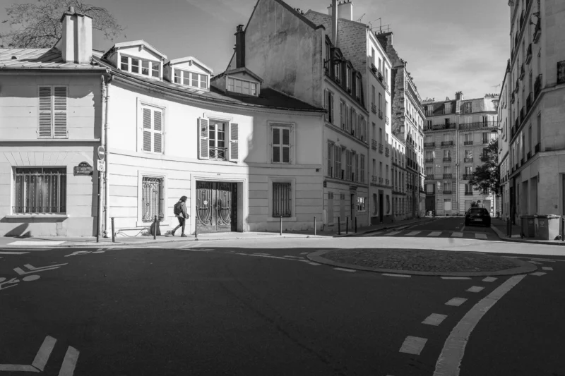Thierry · 10/05/2024 · 3840 x 2560 Cette photographie en noir et blanc capture une rue urbaine calme. Le sujet principal est une intersection peu fréquentée, avec un piéton marchant sur le trottoir. La composition met en valeur des bâtiments classiques aux façades symétriques, typiques d’une architecture européenne.Le cadrage est large, incluant une perspective en profondeur. La lumière, douce et naturelle, crée des contrastes marqués entre ombres et clartés. Les couleurs dominantes, bien que monochromes, jouent sur les nuances de gris.Le style est documentaire et esthétique, évoquant une ambiance paisible et intemporelle, presque mélancolique.