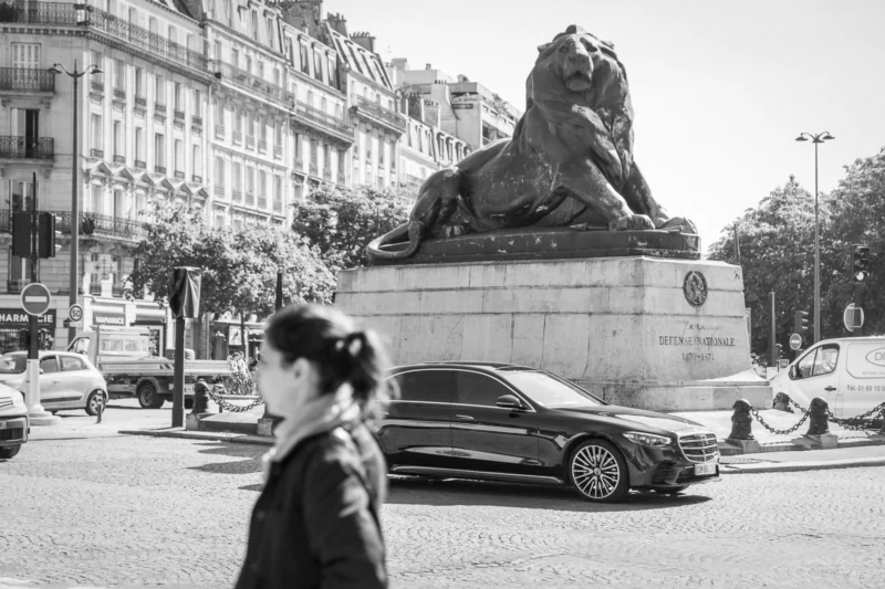 Thierry · 10/05/2024 · 3840 x 2560 Cette photographie en noir et blanc met en scène une femme floue en premier plan, marchant devant une place urbaine. Le sujet principal est la statue imposante d’un lion ou d’un félin sur un piédestal, entourée de voitures et de bâtiments haussmanniens.La composition joue sur les contrastes entre mouvement (la femme) et immobilité (la statue). Le cadrage est serré sur la statue, avec une profondeur de champ réduite. La lumière est douce, typique d’un jour couvert, adoucissant les contours. Les tons dominants sont les gris et noirs, renforçant l’atmosphère à la fois classique et mélancolique. Le style est documentaire et intemporel, évoquant une ambiance parisienne paisible et légèrement nostalgique.