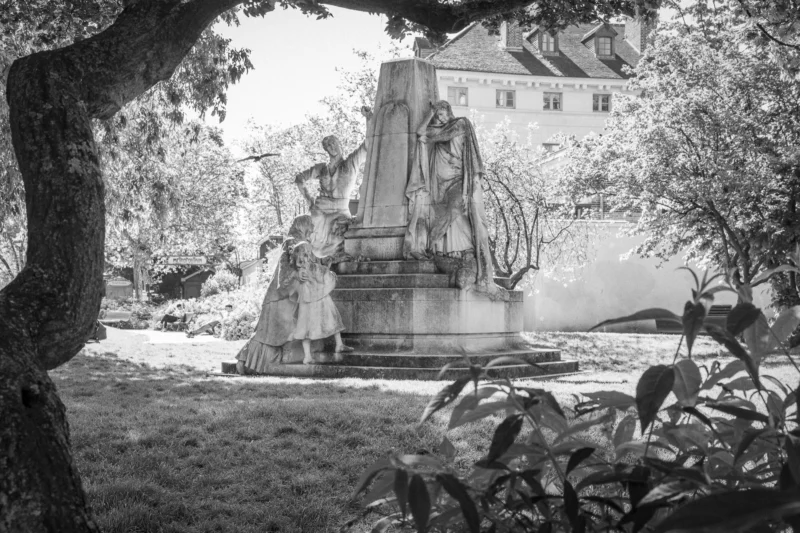 Thierry · 10/05/2024 · 3840 x 2560 Cette photographie en noir et blanc met en scène un monument funéraire comme sujet principal, représentant une figure féminine en deuil. La composition montre une statue imposante entourée de végétation, avec un cadrage serré qui accentue les détails et l'atmosphère solennelle.La lumière douce, probablement naturelle, crée des contrastes et des ombres marquées. Les couleurs dominantes, bien que monochromes, exploitent les nuances de gris pour renforcer le côté dramatique. Le style est classique et mélancolique, évoquant une ambiance historique et nostalgique, renforcée par le flou des feuilles en premier plan.