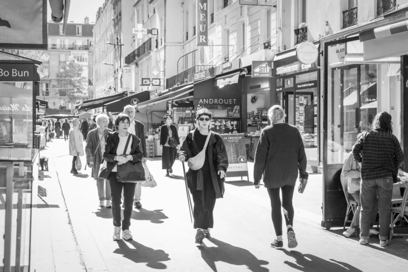 Thierry · 10/05/2024 · 3840 x 2560 Cette photographie en noir et blanc capture une rue animée en ville. Le sujet principal est un groupe de piétons marchant sur un trottoir étroit, entourés de commerces.La composition est dynamique, avec une perspective légèrement en contre-plongée. Le cadrage inclut des bâtiments et des enseignes de part et d'autre, créant une atmosphère urbaine dense.La lumière est naturelle, douce et diffuse, typique d’un jour nuageux. Les contrastes sont modérés, accentuant les détails des visages et des textures.Le style est documentaire et réaliste, évoquant une ambiance quotidienne et authentique dans un cadre urbain.