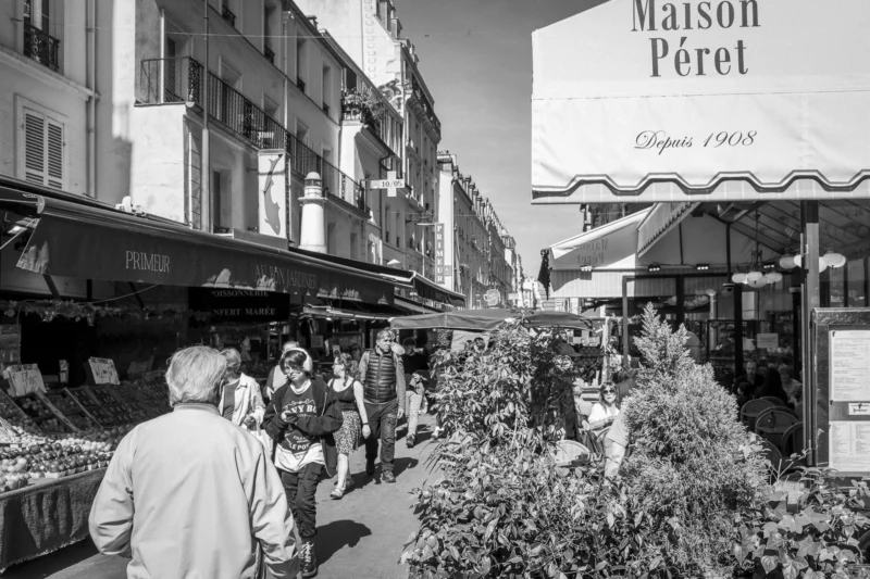 Thierry · 10/05/2024 · 3840 x 2560 Cette photographie en noir et blanc capture une scène de marché animé. Le sujet principal est un groupe de personnes dans une rue commerçante, devant des étals de fleurs et des boutiques.La composition montre une perspective légèrement en contre-plongée, mettant en valeur les passants et les enseignes. Le cadrage est serré sur les détails des stands et les visages floutés des gens.La lumière est naturelle et semble provenir d’un jour ensoleillé, créant des contrastes marqués. Les couleurs dominantes, bien que monochromes, évoquent des tons gris et noirs avec quelques touches de blanc.Le style est documentaire et réaliste, évoquant une ambiance vivante et typique d’un marché urbain.