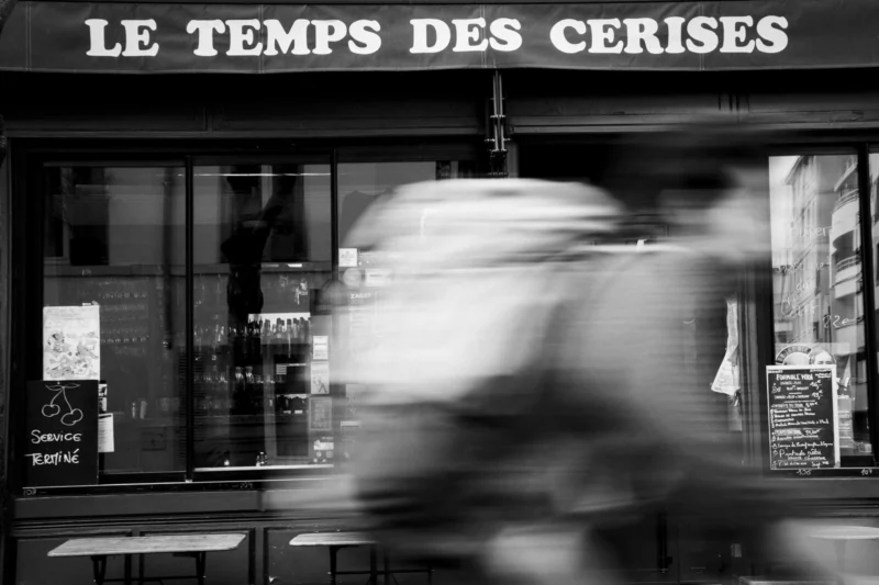 Thierry · 28/03/2024 · 3840 x 2560 Cette photographie en noir et blanc capture une scène floue d’une personne en mouvement devant un café nommé **« Le Temps des Cerises »**. Le sujet principal est le flou dynamique de la silhouette, contrastant avec les éléments nets de l’établissement.La composition joue sur le contraste entre le flou artistique et les détails précis du café (enseigne, vitrine, chaises). Le cadrage est serré, centré sur l’entrée du commerce. La lumière est douce, créant des ombres marquées et une ambiance nocturne ou crépusculaire. Les tons dominants sont les gris et noirs, renforçant une atmosphère à la fois moderne et rétro. Le style évoque un mélange de street photography et de flou intentionnel pour suggérer le mouvement.