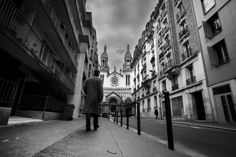 Thierry · 04/05/2024 · 3840 x 2560 Cette photographie en noir et blanc met en scène une rue urbaine étroite avec un homme en imperméable comme sujet principal, marchant vers une cathédrale gothique imposante en arrière-plan.La composition utilise une perspective convergente, accentuant la profondeur. Le cadrage est serré, encadrant les bâtiments de chaque côté. La lumière est tamisée, créant des contrastes marqués et une ambiance mélancolique. Les tons dominants sont les gris et noirs, renforçant l'atmosphère sombre et contemplative. Le style rappelle le photojournalisme classique, évoquant une ambiance à la fois mystérieuse et introspective.