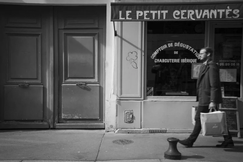 Thierry · 08/03/2024 · 3840 x 2560 Cette photographie en noir et blanc met en scène un homme marchant devant une boutique nommée **« Le Petit Cervantès »**, spécialisée en déshabillage et charpenterie génésique.Le sujet principal est l’homme, vêtu d’un manteau et tenant une mallette. La composition est équilibrée, avec un cadrage serré sur la porte et l’entrée. La lumière, tamisée, crée des contrastes doux, accentuant les ombres et les détails architecturaux. L’ambiance est à la fois urbaine et intime, évoquant une atmosphère nocturne mystérieuse et légèrement rétro. Le style est documentaire, avec une touche artistique.